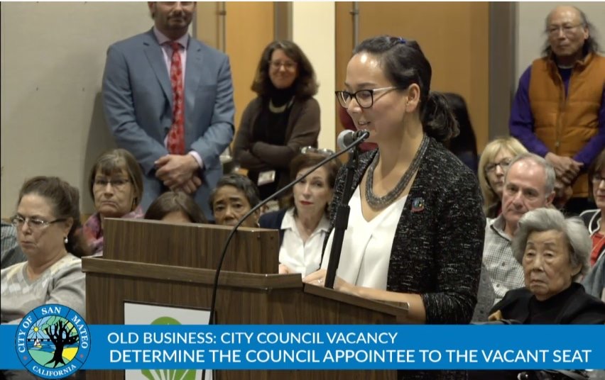 Amourence Lee standing at a podium in front of a crowd interviewing for the open city council appointment.